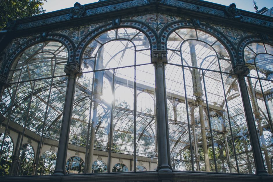 Close-up view of a large, ornate glass greenhouse with curved, arched windows and intricate metal framing, set against a clear blue sky. The structure features multiple glass panels, some reflecting s