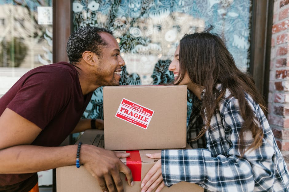 A man and a woman are outdoors in front of a building with a large window. They are smiling and leaning close to each other, holding or sharing two cardboard boxes stacked on top of each other. The to