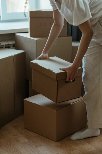 A man and a woman are outdoors in front of a building with a large window. They are smiling and leaning close to each other, holding or sharing two cardboard boxes stacked on top of each other. The to
